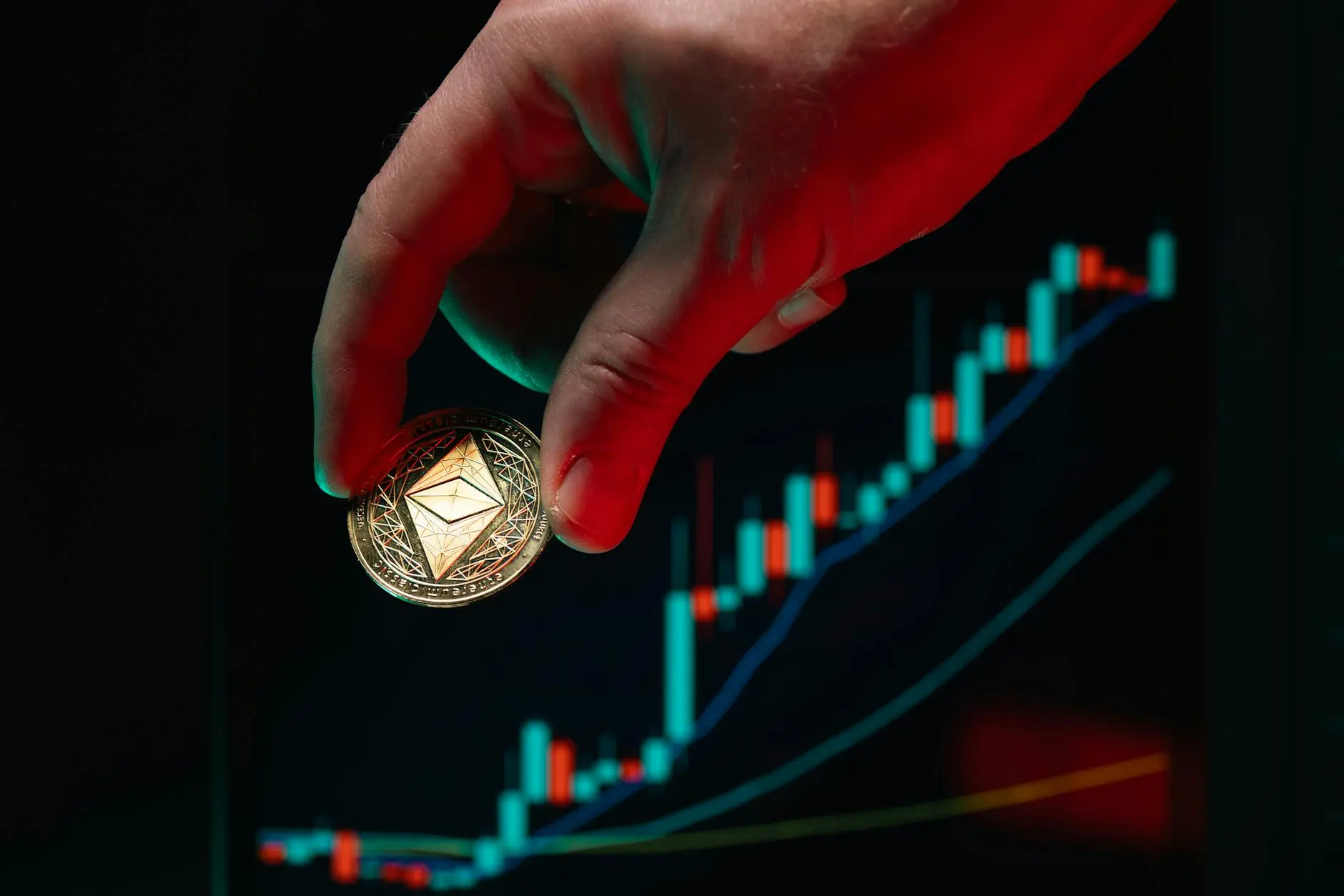 A hand holding a physical Ethereum coin in front of a trading chart.